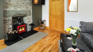 A living room with a stone fireplace, a flat-screen TV, a wooden door, a grey sofa, and a table with a vase of flowers. - 1 bedroom accommodation in Dunoon (Dunoon)