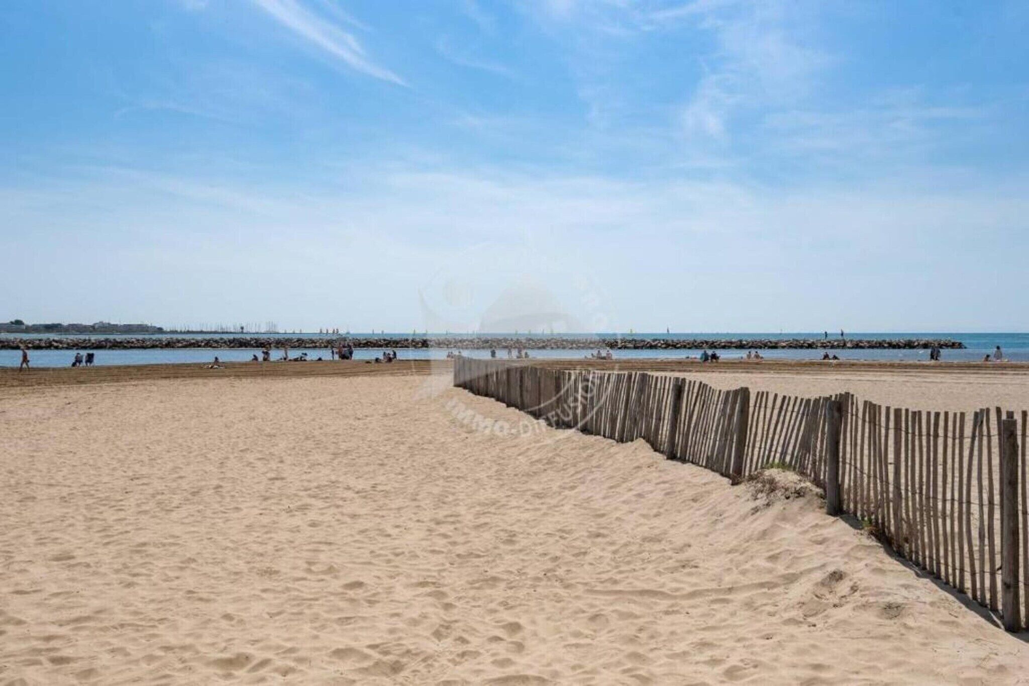 Een zandstrand met een houten omheining op de voorgrond en een steiger in de verte.