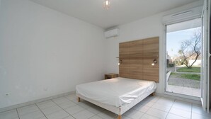 Une chambre à coucher avec un lit, une tête de lit en bois, une petite table de chevet et une grande fenêtre donnant sur l’extérieur.