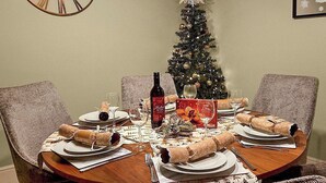 Ein Esstisch ist für vier Personen gedeckt, mit Tellern, Gläsern und einer Weinflasche. Im Hintergrund steht ein Weihnachtsbaum mit Dekorationen.