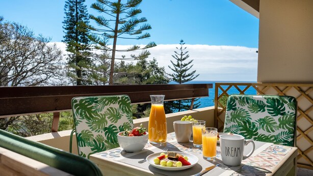 A balcony with a table set for breakfast, including a bowl of fruit, a bottle of orange juice, and a mug with the word "DREAM" on it. - Solar do Mar by LovelyStay (Funchal)