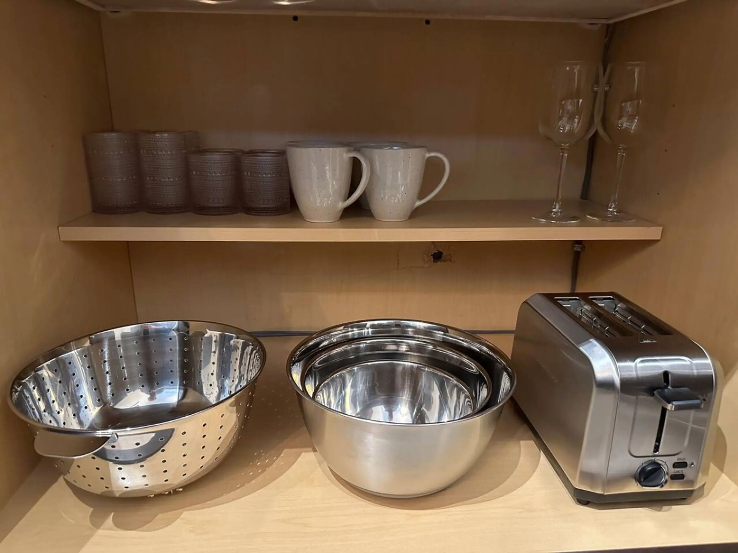 A kitchen shelf with a toaster, two stainless steel colanders, and a row of white and brown cups and glasses.