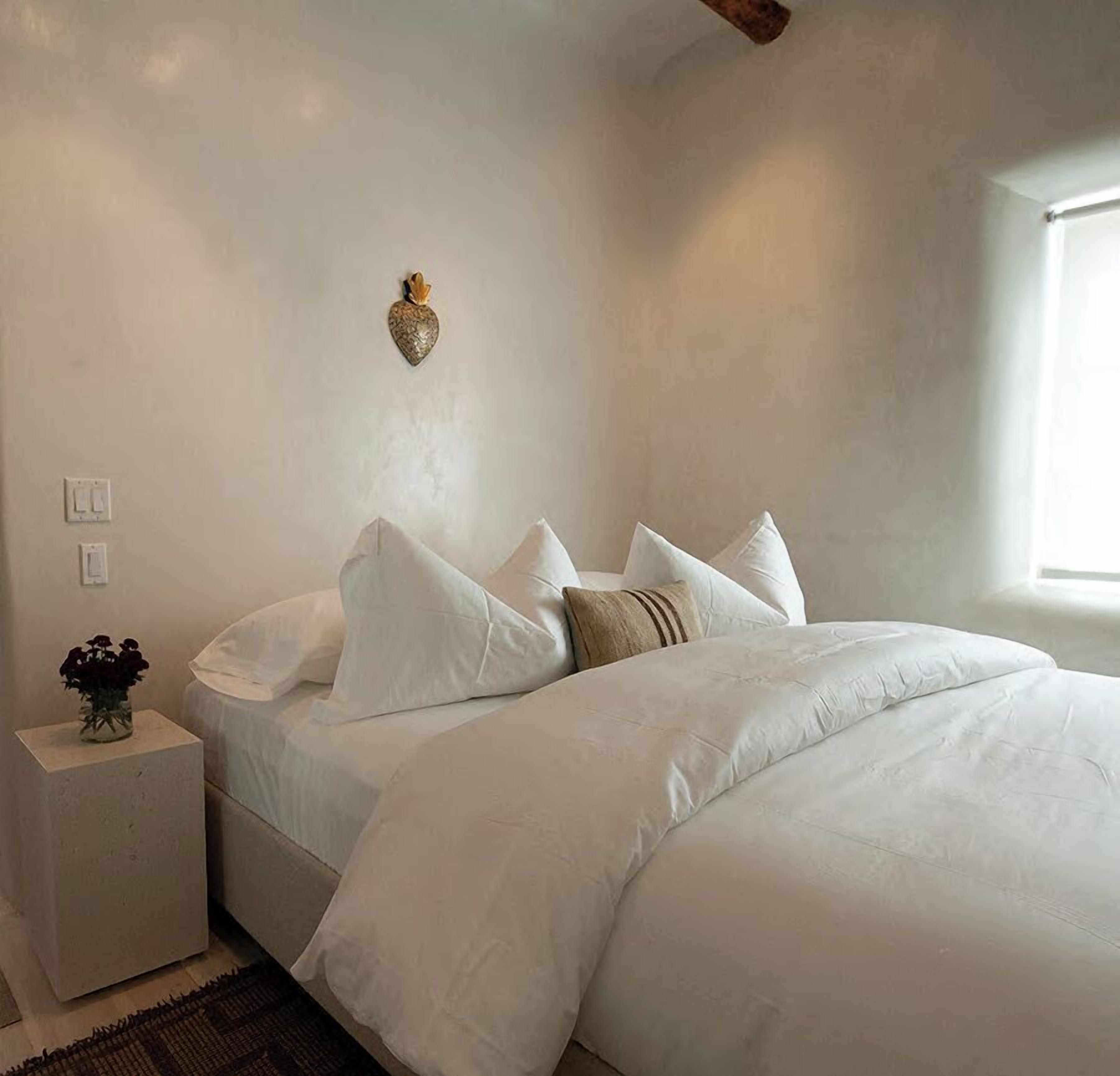 A bedroom with a bed, white walls, a small bedside table with a vase of flowers, and a window with natural light.