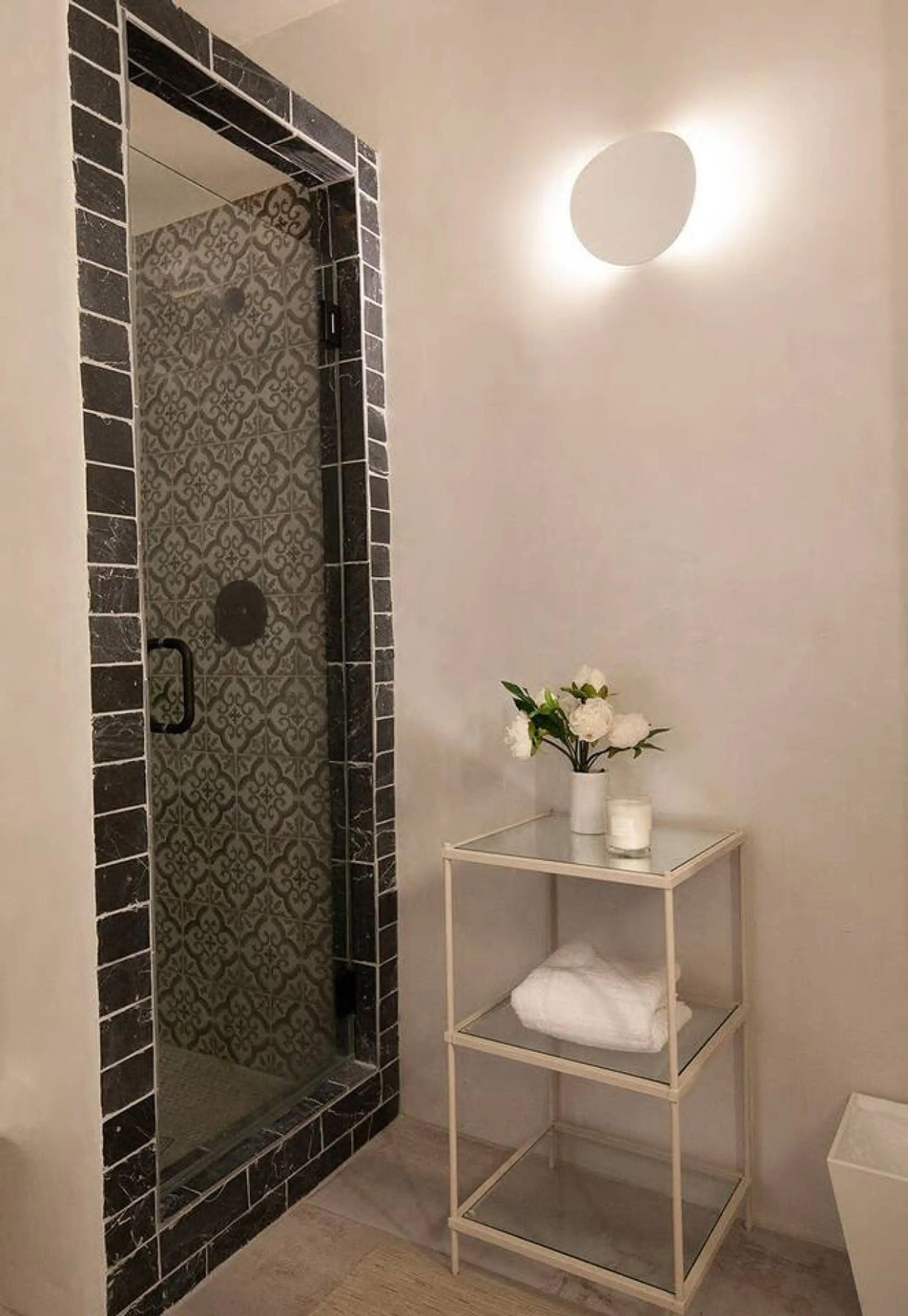 Un baño con ducha revestida de azulejos negros, un estante de vidrio con un florero y una lámpara de pared redonda.