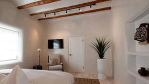 Un dormitori amb un llit, una cadira, una planta en test, un televisor i una estant amb objectes decoratius.
