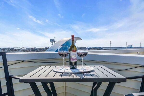 A table with a bottle and two glasses of red wine on a balcony overlooking a body of water and a distant shoreline.