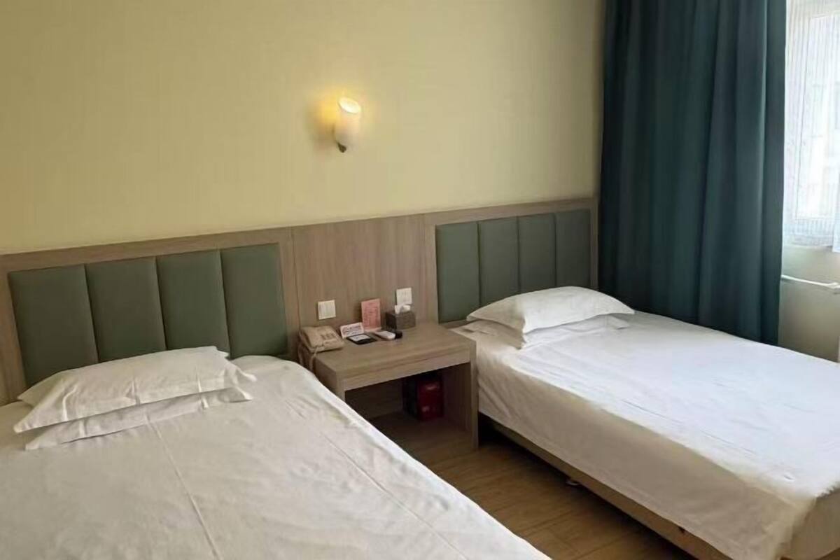 A hotel room with two single beds, a bedside table with a phone and a lamp, and a window with curtains.