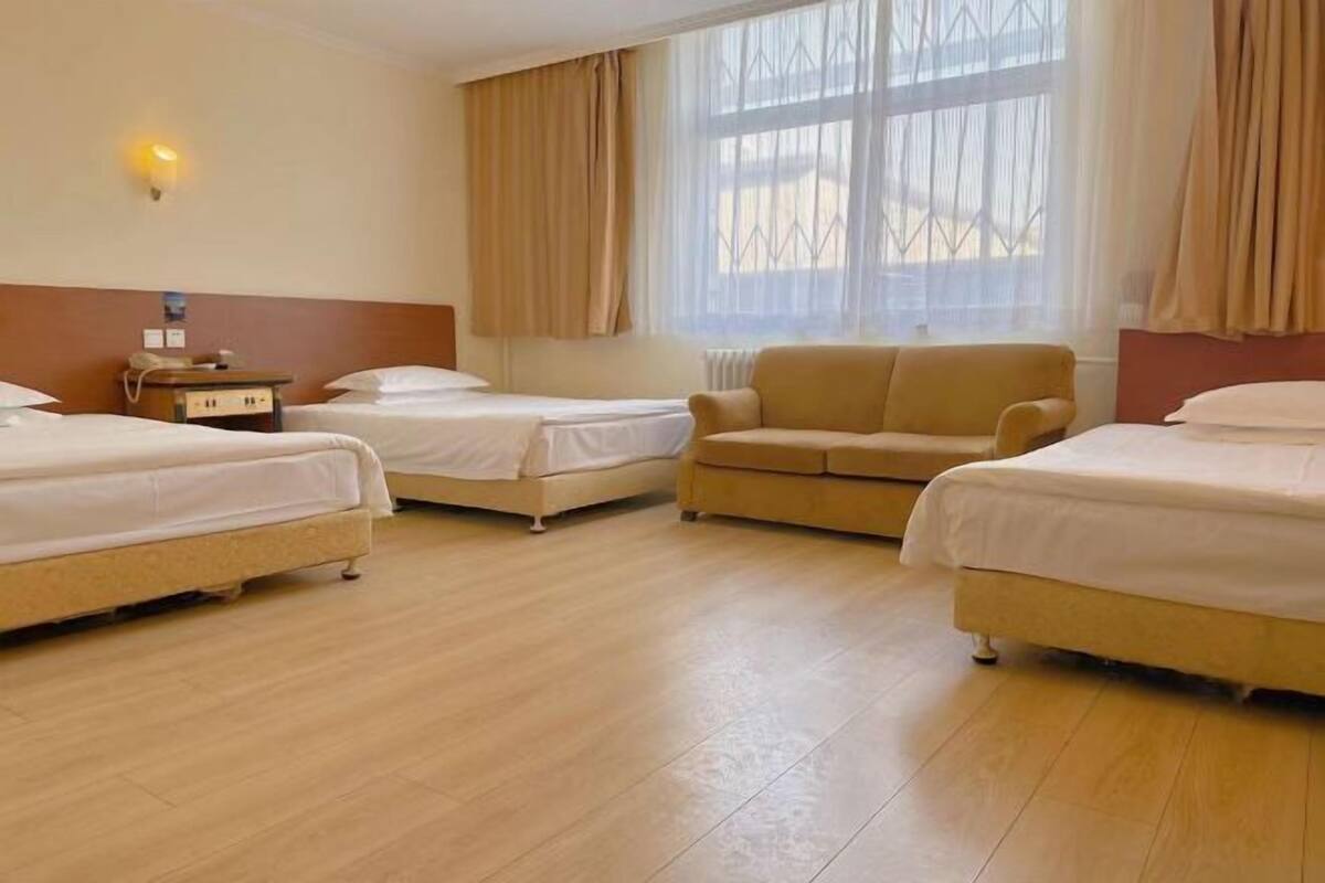 A hotel room with two single beds, a sofa, a bedside table with a lamp, and a window with curtains.