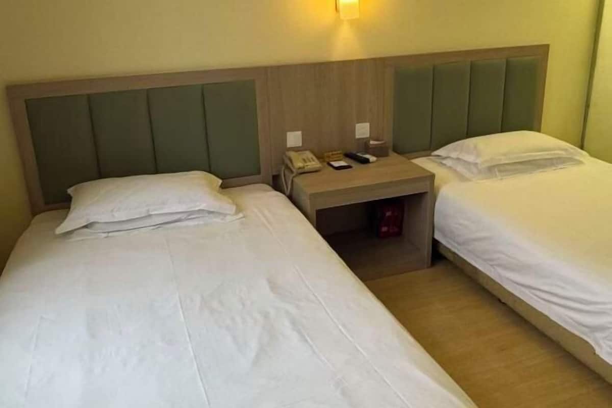 A hotel room with two beds, a wooden headboard, a nightstand with a phone and remote, and a wall-mounted light.