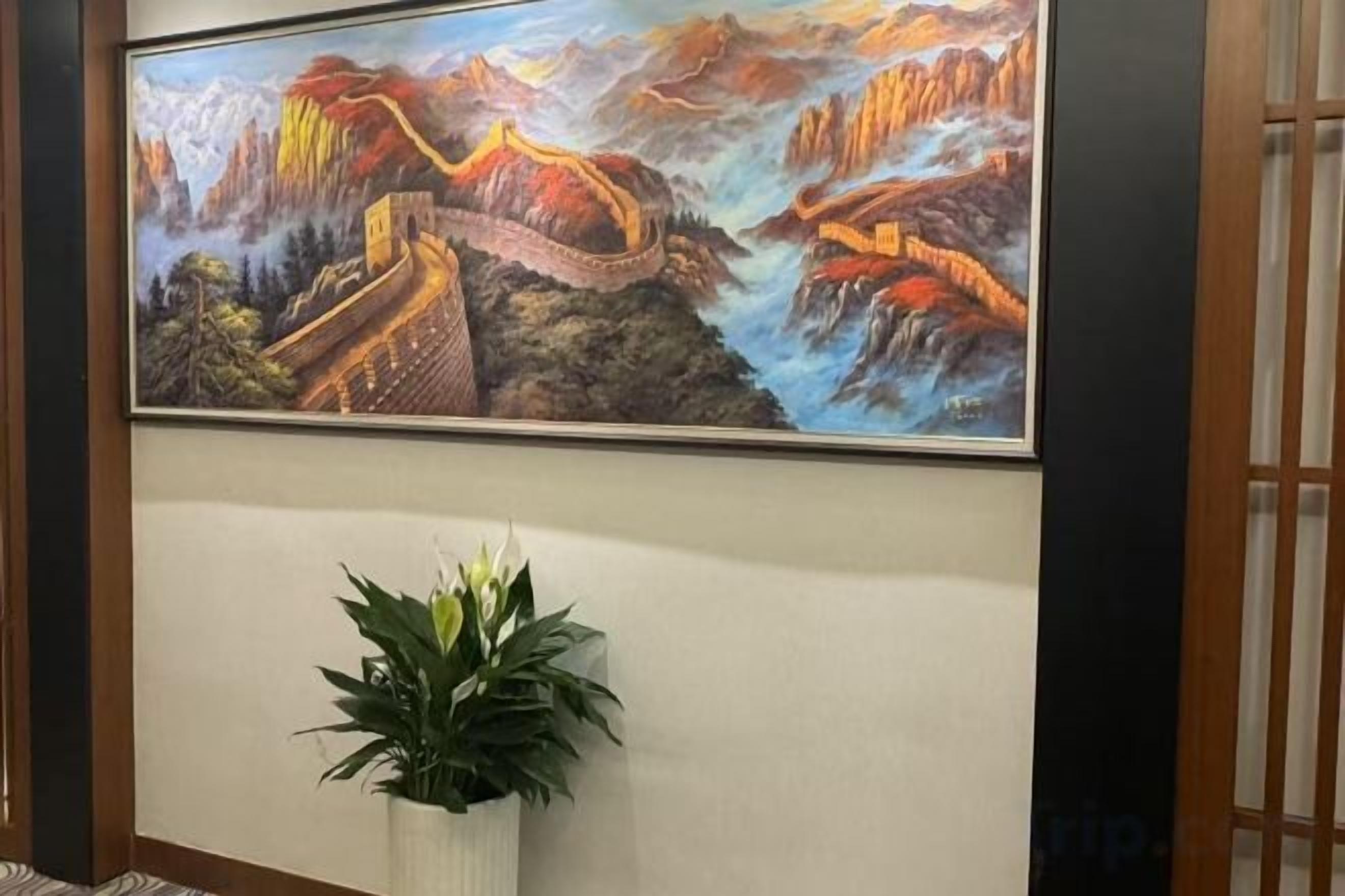 Un cuadro enmarco de la Gran Muralla China sobre una pared, una planta en maceta y una puerta de madera con listones verticales.