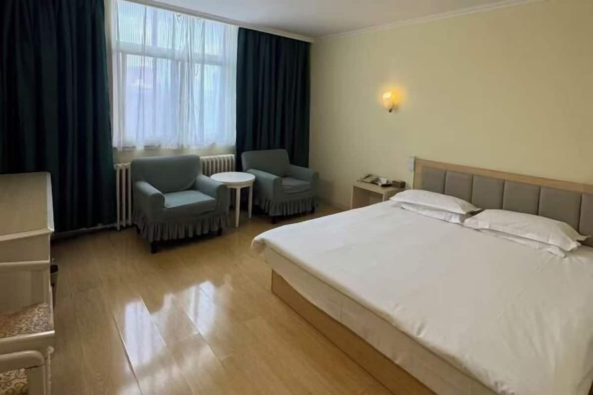 A hotel room with a large bed, two armchairs, a small table, a nightstand, and a window with curtains.