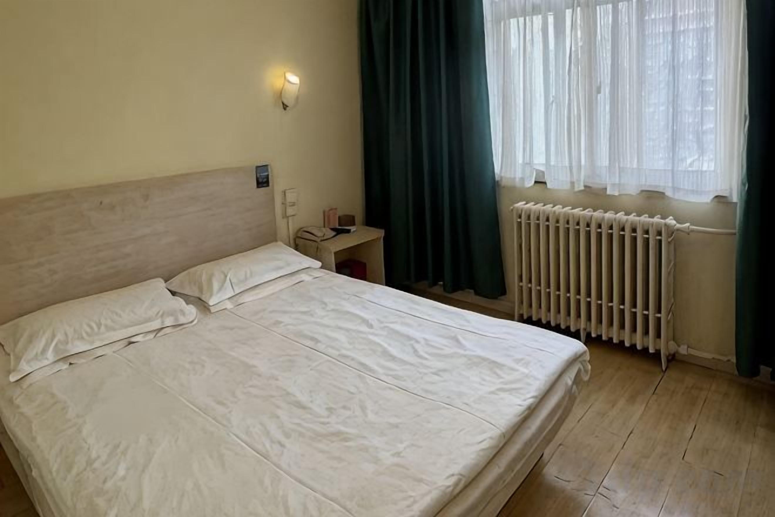 Ein Hotelzimmer mit Bett, Nachttisch, Lampe, einem Fenster mit Vorhängen und einem Heizkörper.