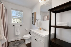A modern bathroom with a white toilet, a large window, a white sink with a mirror above it, and a wooden shelf with towels. - Spacious & Updated 4-bedroom home w/BBQ, fire pit & AC in the Heart of Missoula! (Missoula)