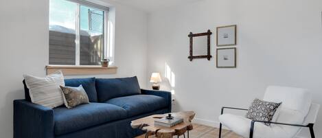 Una sala de estar con un sofá azul, un sillón blanco, una mesa de centro de madera y una mesita auxiliar.