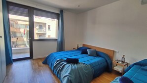 A bedroom with two beds, a window with blinds, a wooden floor, and a small bedside table with a lamp. - Elegant Apartment with Terrace – A Peaceful Oasis in Palermo (Palermo)