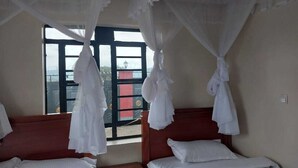 Ein Zimmer mit zwei Betten, jeweils mit weißer Bettwäsche und einem hölzernen Kopfteil. Weiße Vorhänge hängen von der Decke, und ein Fenster bietet Blick auf ein Gebäude.