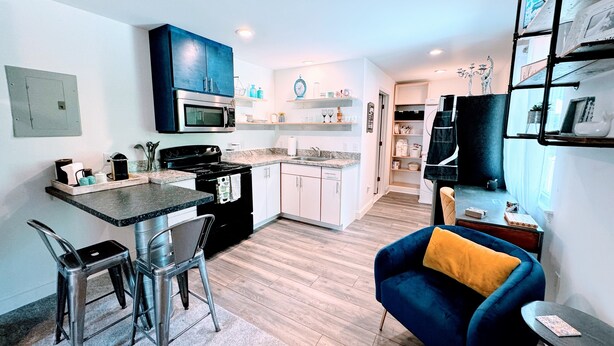 A modern kitchen with a microwave, stove, and bar stools. There is a blue armchair with a yellow cushion in the living area. - “See you by the wall” Duke region (Durham)