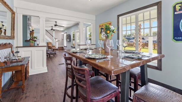 A dining area with a wooden table set for six, surrounded by wooden chairs. A large window with a view of the outdoors is visible.