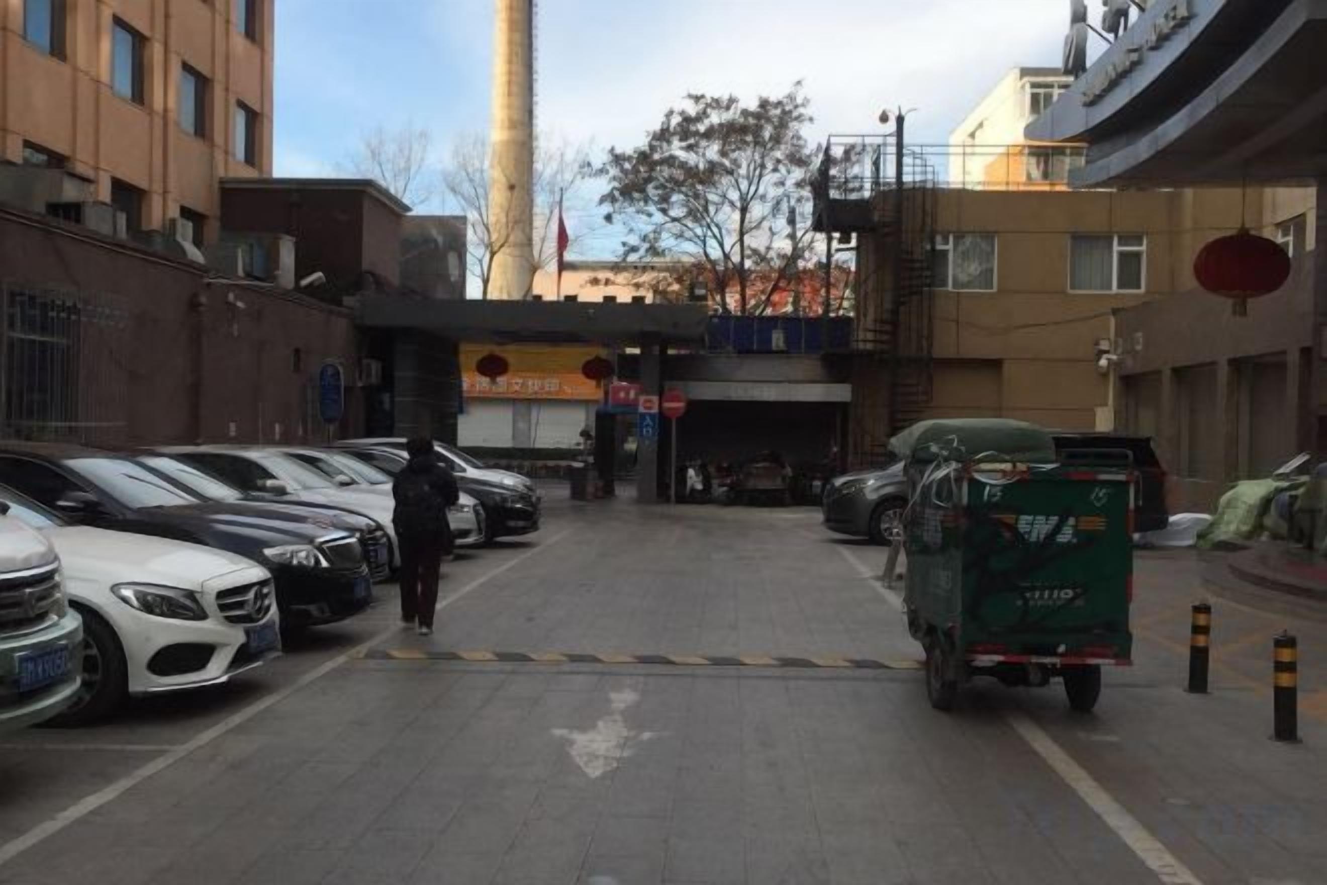 A parking lot with cars and a green truck, buildings in the background, and a clear sky.