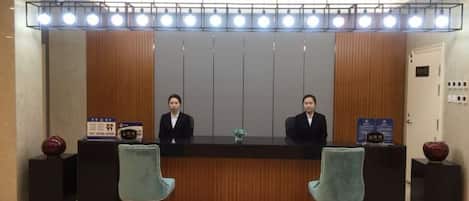 A reception desk with two staff members, a wooden panel backdrop, and a ceiling light fixture.