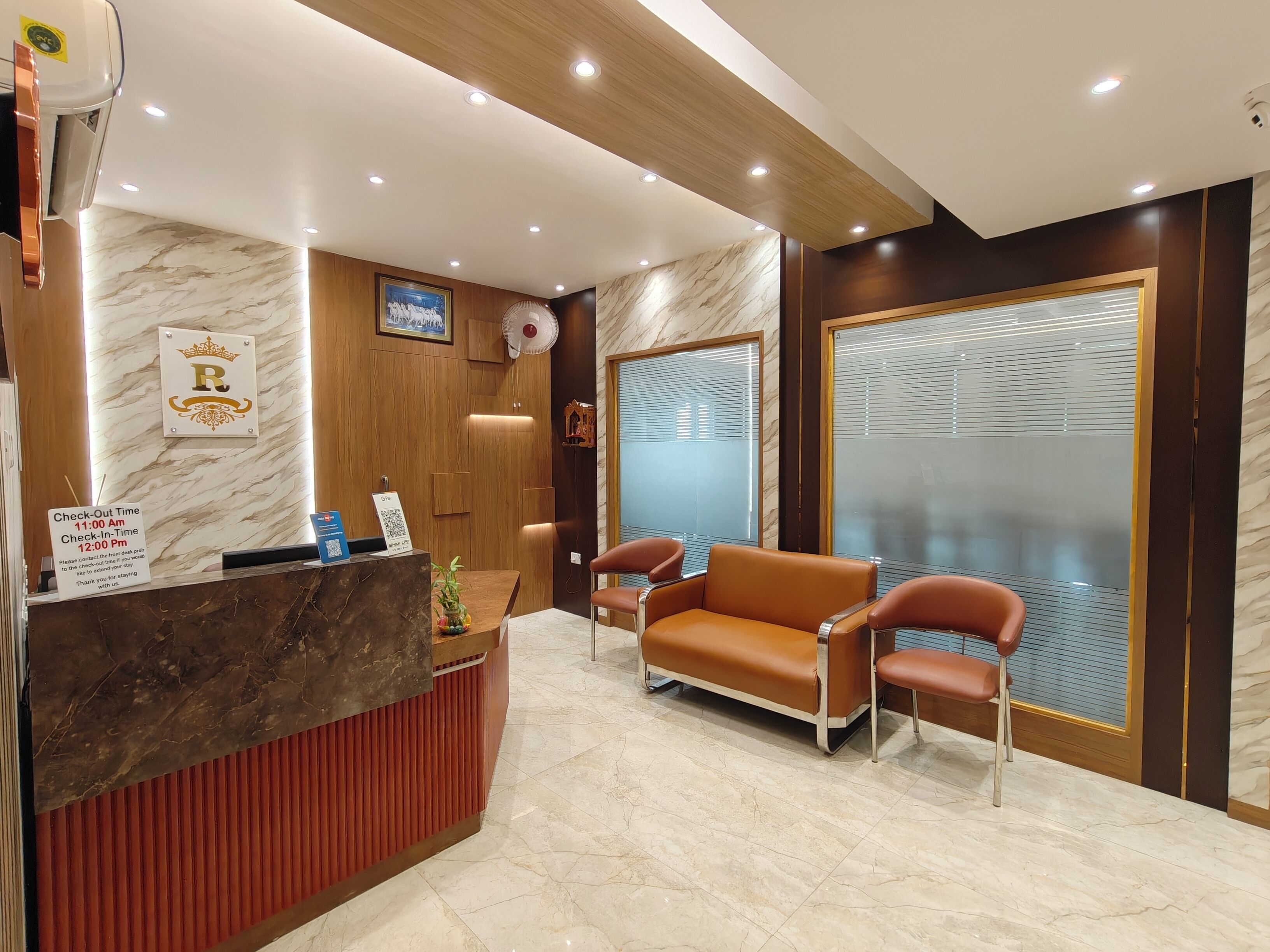 Zona de recepción de un hotel con piso de mármol, mostrador de recepción, cartel de salida y un área de estar con sillones de cuero, además de un ventanal con persianas.