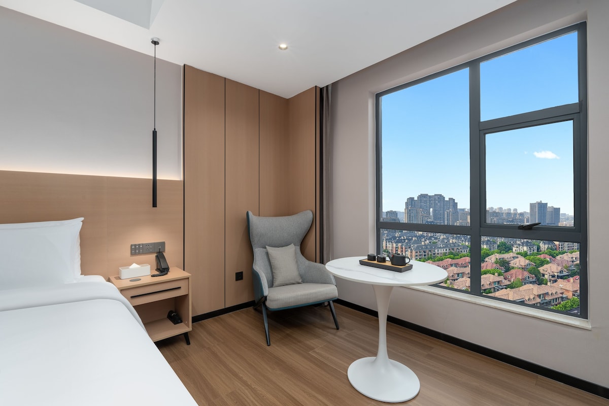 A modern hotel room with a bed, a nightstand, a chair, a small table, and a large window offering a city view.