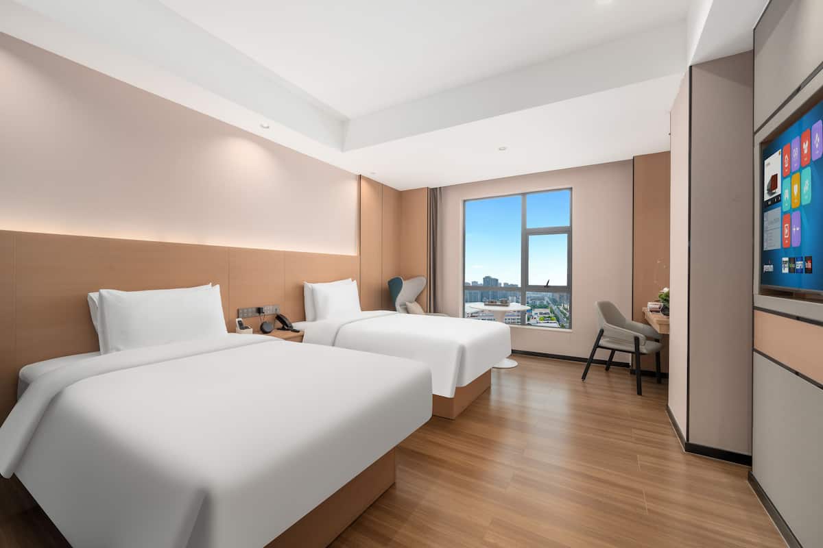 Ett hotellrum med två sängar, ett skrivbord, en stol, en TV och ett stort fönster med utsikt över staden.