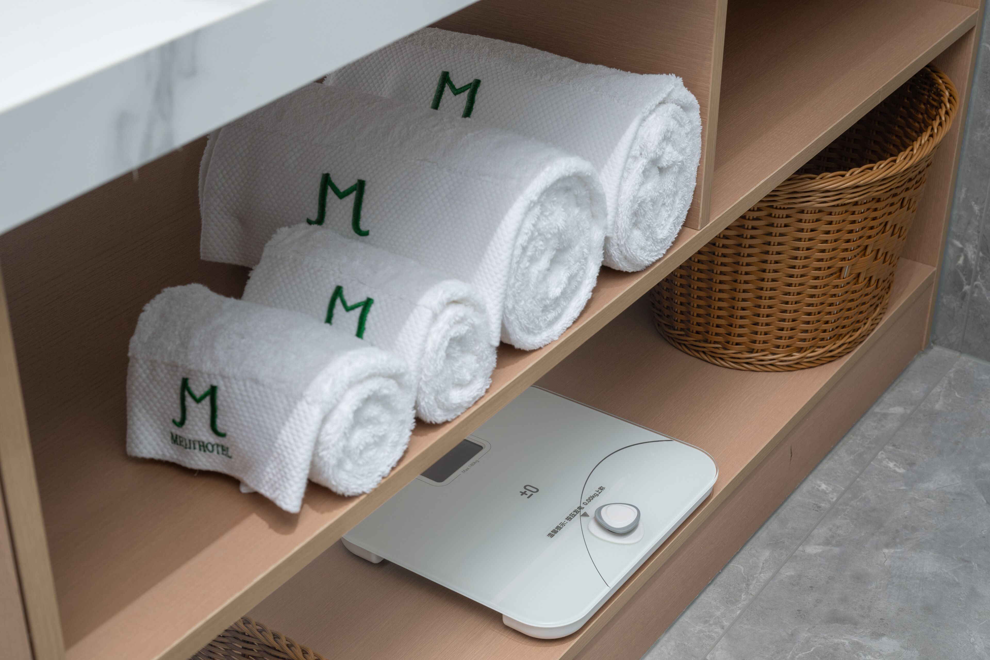 A bathroom shelf with neatly folded towels, a digital scale, and a wicker basket.