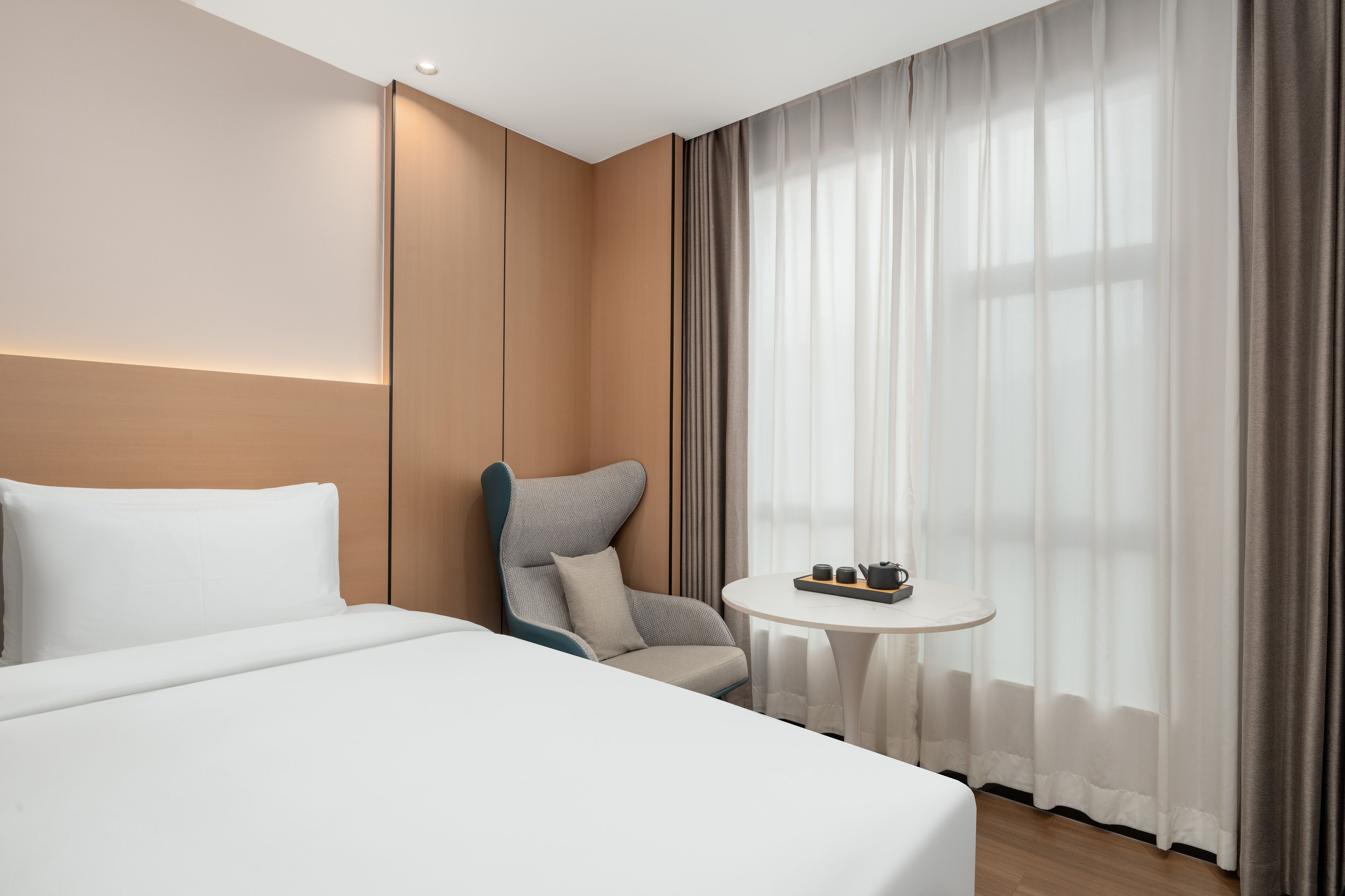 A hotel room with a large bed, a small round table with a tray of items, a chair, and a window with curtains.