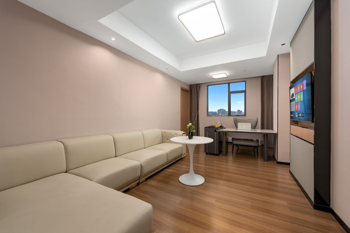 A modern hotel room with a sofa, a small round table, a desk with a laptop, a TV, and a city view.