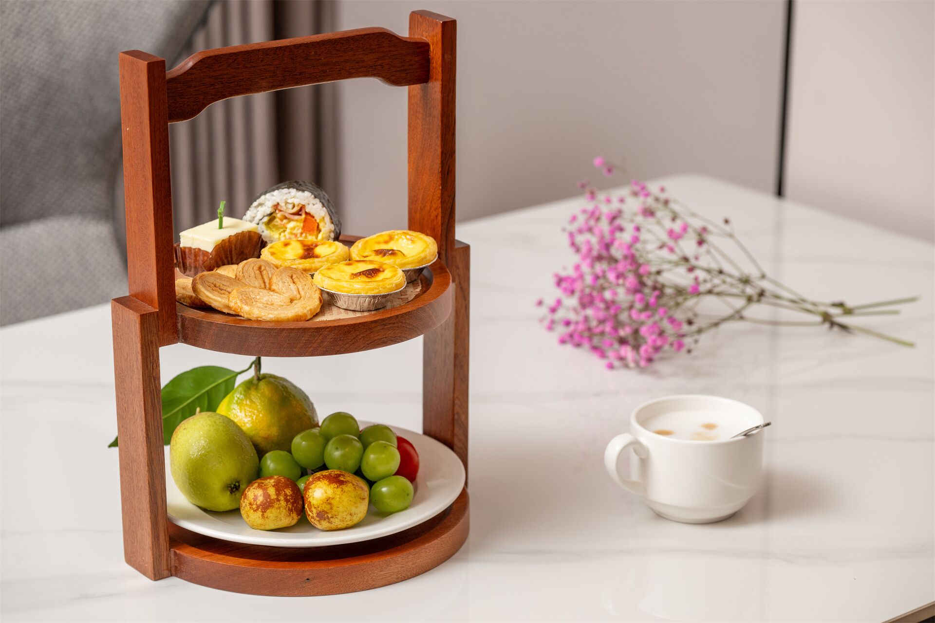 A wooden three-tiered serving tray with pastries, fruits, and a cup of coffee on a white table.