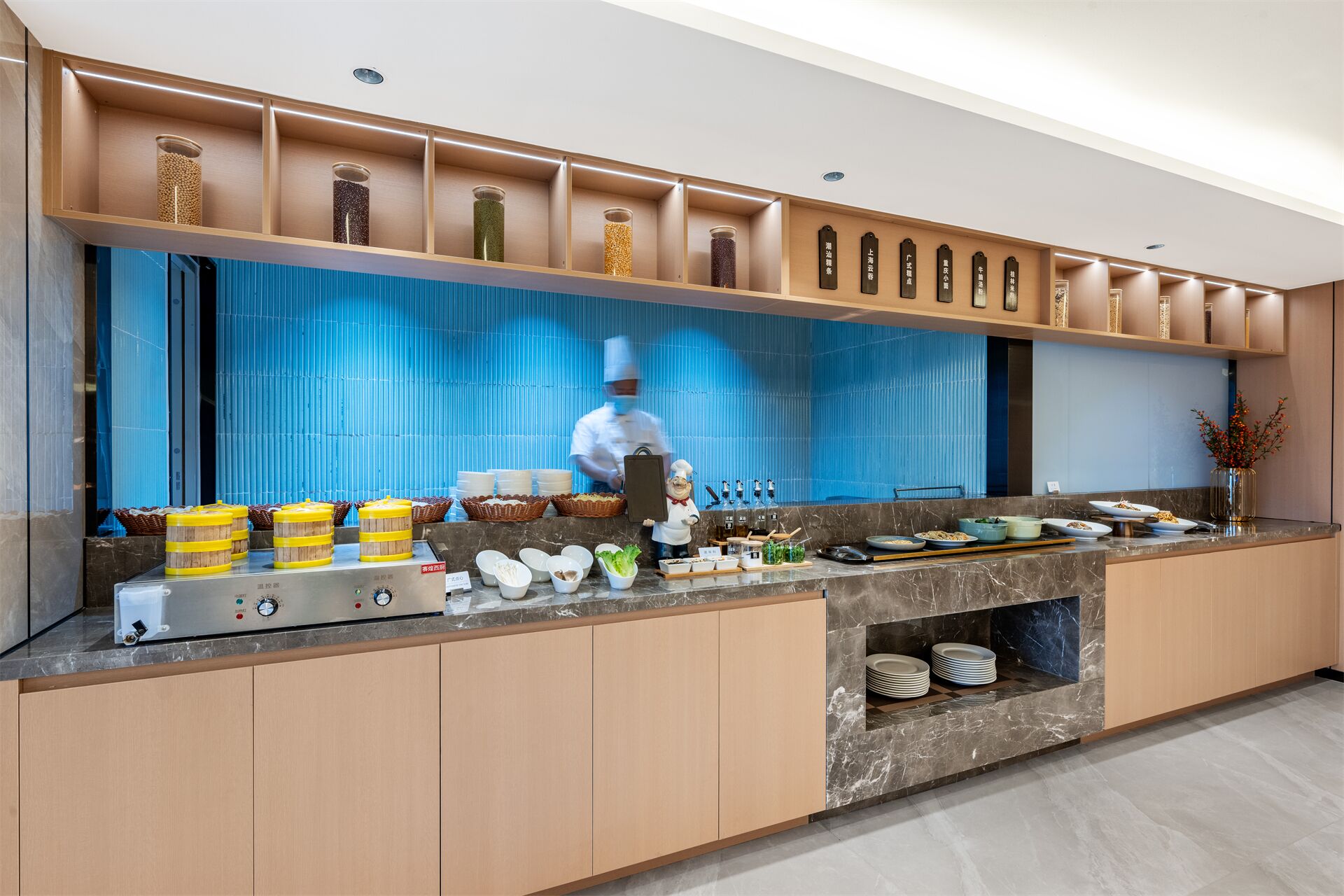 Une cuisine moderne où un chef prépare des plats. On y voit un mur de couleur bleue derrière le plan de travail, des armoires en bois et divers ingrédients disposés sur le comptoir.