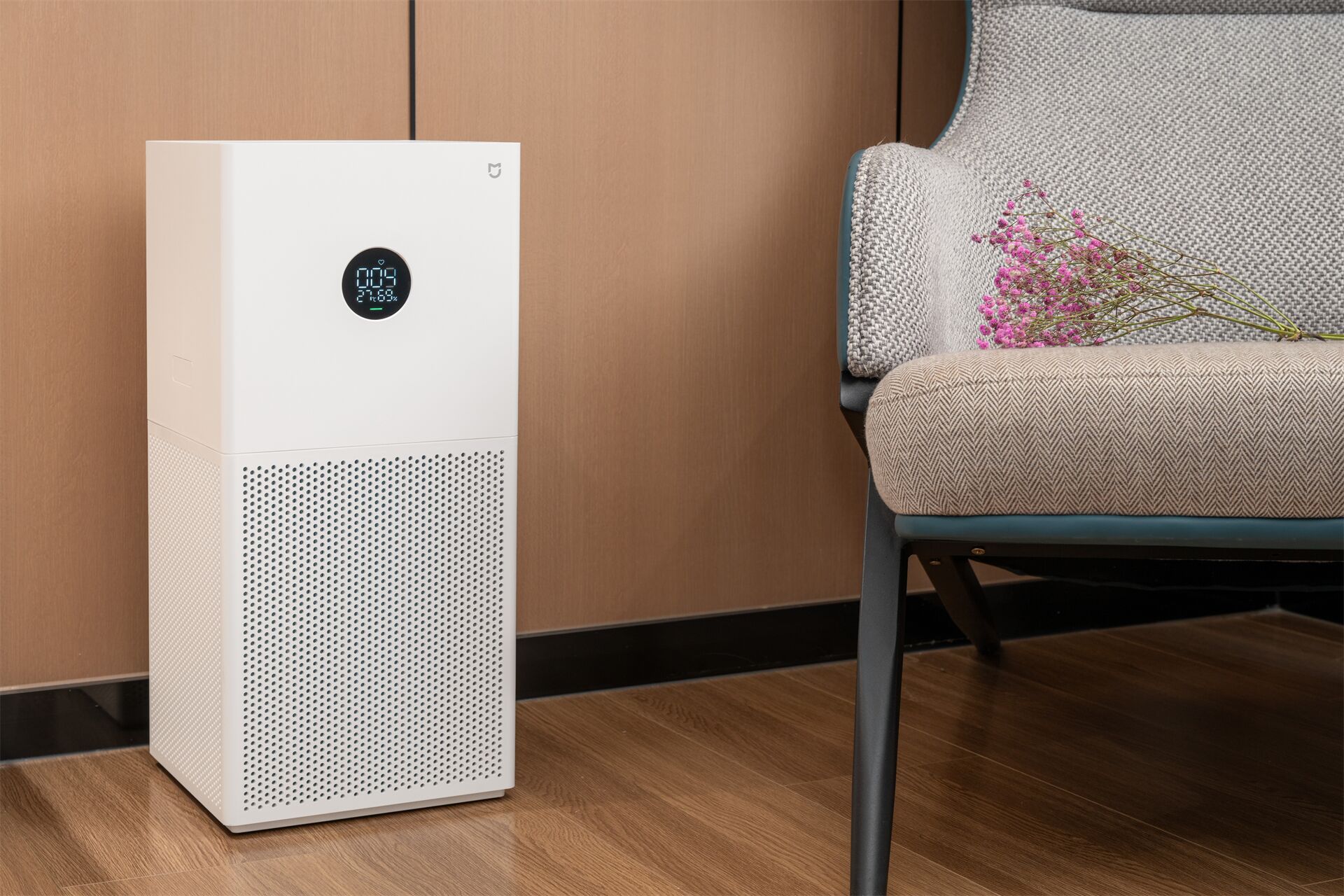 A white air purifier with a digital display, placed next to a chair with a woven cushion and a bouquet of flowers.