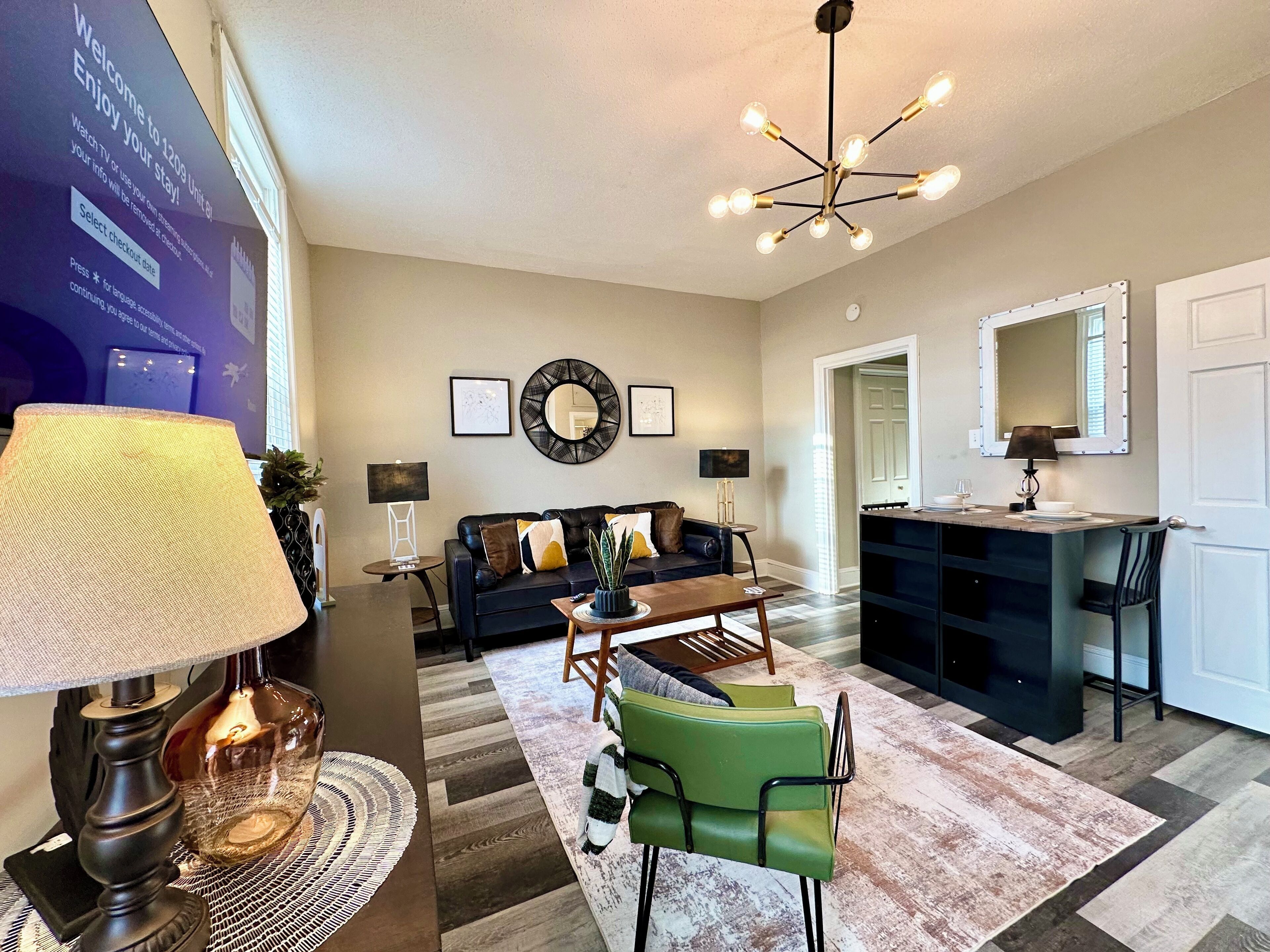 A modern living room with a black leather sofa, a coffee table, and a green armchair. There is a chandelier with multiple bulbs, a wall-mounted mirror, and framed artwork.