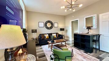 A modern living room with a black leather sofa, a coffee table, and a green armchair. There is a chandelier with multiple bulbs, a wall-mounted mirror, and framed artwork.