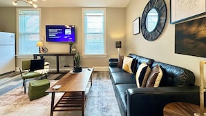 A living room with a black leather sofa, a wooden coffee table, a TV on the wall, and a large mirror.