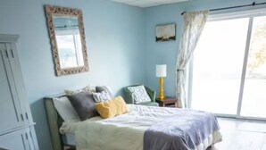 A bedroom with a wooden floor, a bed with a white and gray duvet, a green armchair, a lamp, and a window with curtains. - Panoramic Picturesque Home in Blue Ridge Mtns (Max Meadows)