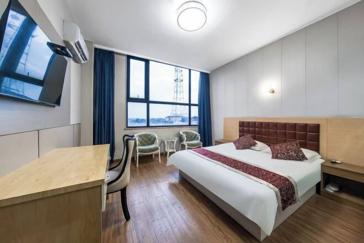 A hotel room with a large bed, a desk, two chairs, a TV, and a window with a city view.