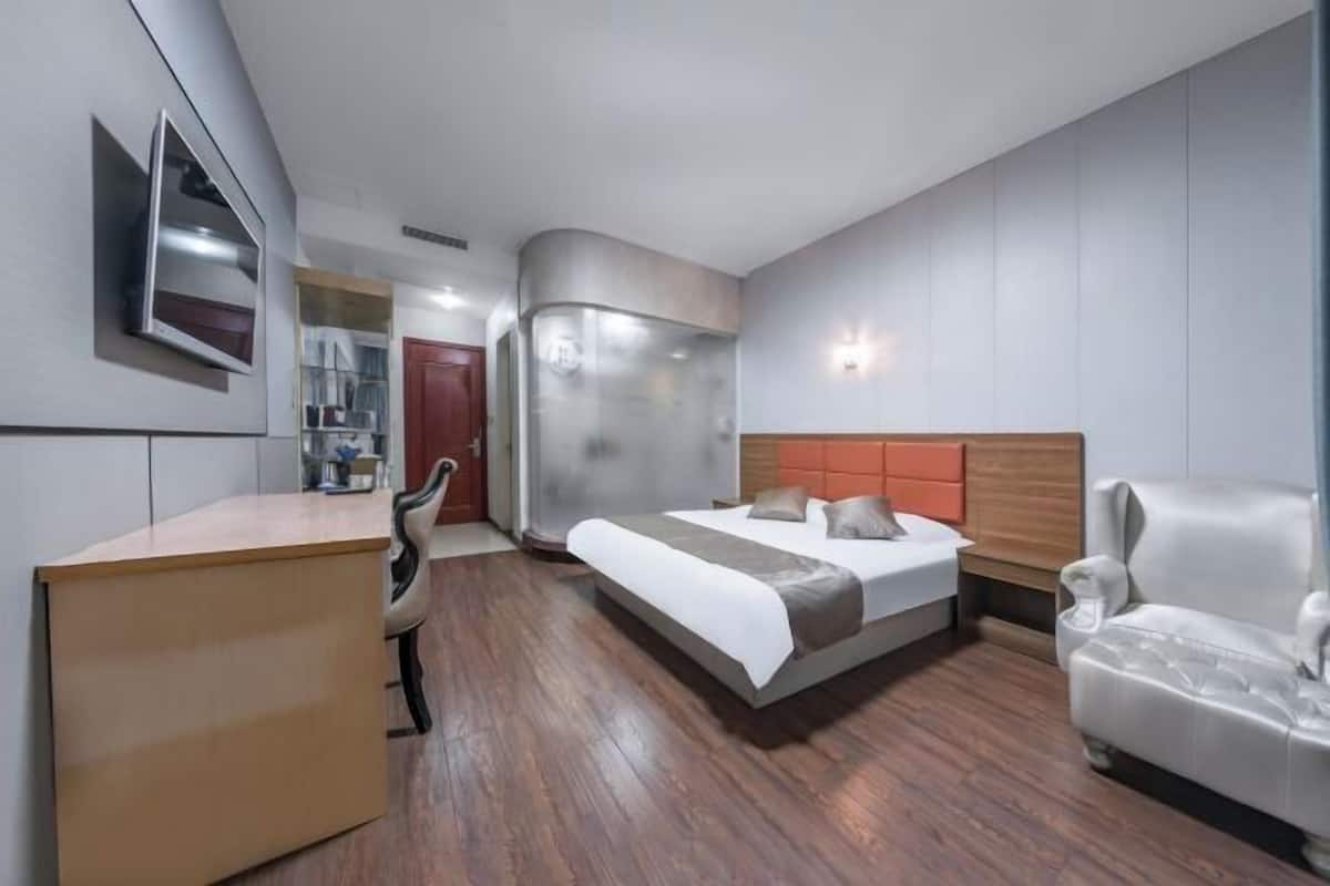 A modern hotel room with a wooden desk, a bed with a grey and white headboard, and a white armchair.