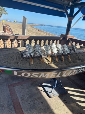 A beachside grill with grilled fish, a wooden fence, and a clear view of the beach and ocean. - Apartment With sea Views in Benalbeach (Benalmádena)