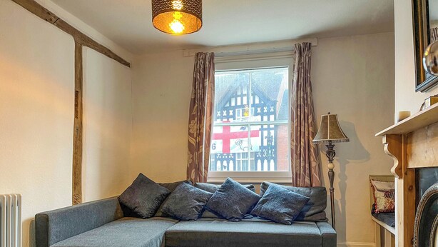 Ein gemütlicher Wohnraum mit einem grauen Sofa, einem Couchtisch mit Zeitschriften, einem Kamin und einem Fenster mit Blick auf ein Fachwerkhäuschen.