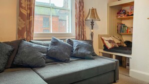Ein gemütliches Wohnzimmer mit einer grauen Couch, gemusterten Vorhängen, einer Lampe und einem Bücherregal.
