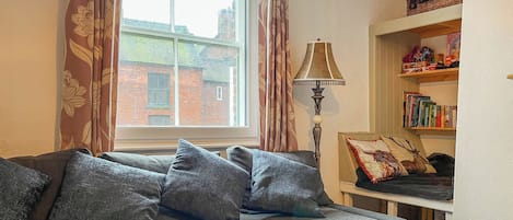 Una sala acogedora con un sofá gris, cortinas estampadas, una lámpara y una estantería.