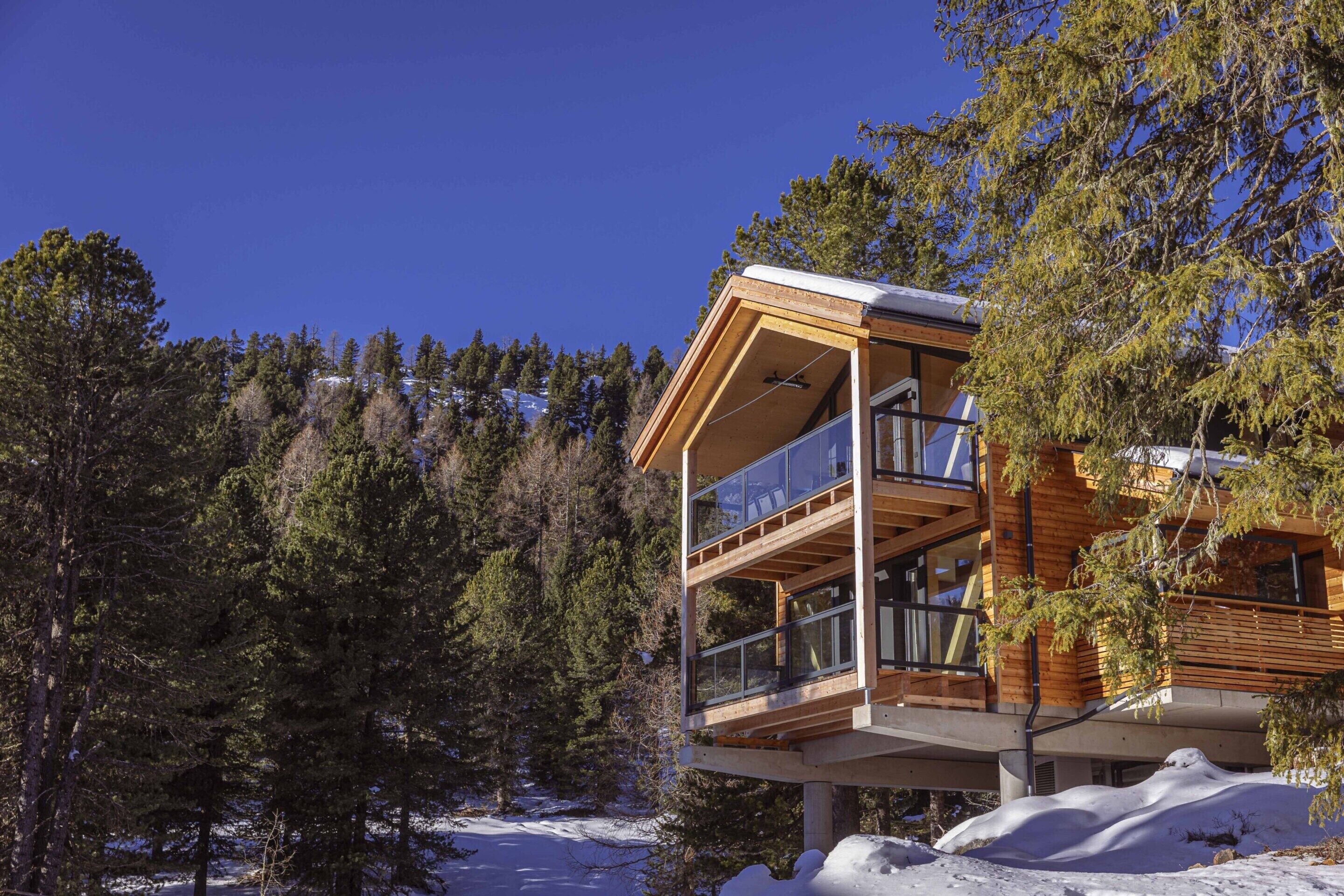 A modern wooden cabin with a glass balcony, surrounded by snow and trees.
