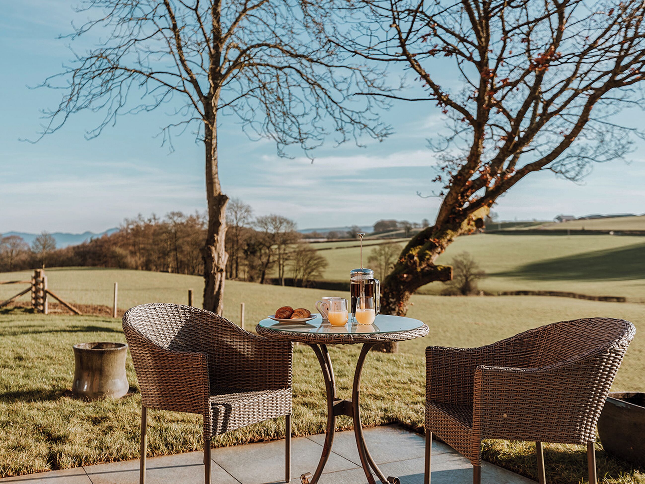 A wicker table with two chairs and a pitcher of juice, set on a patio with a view of rolling hills and trees.