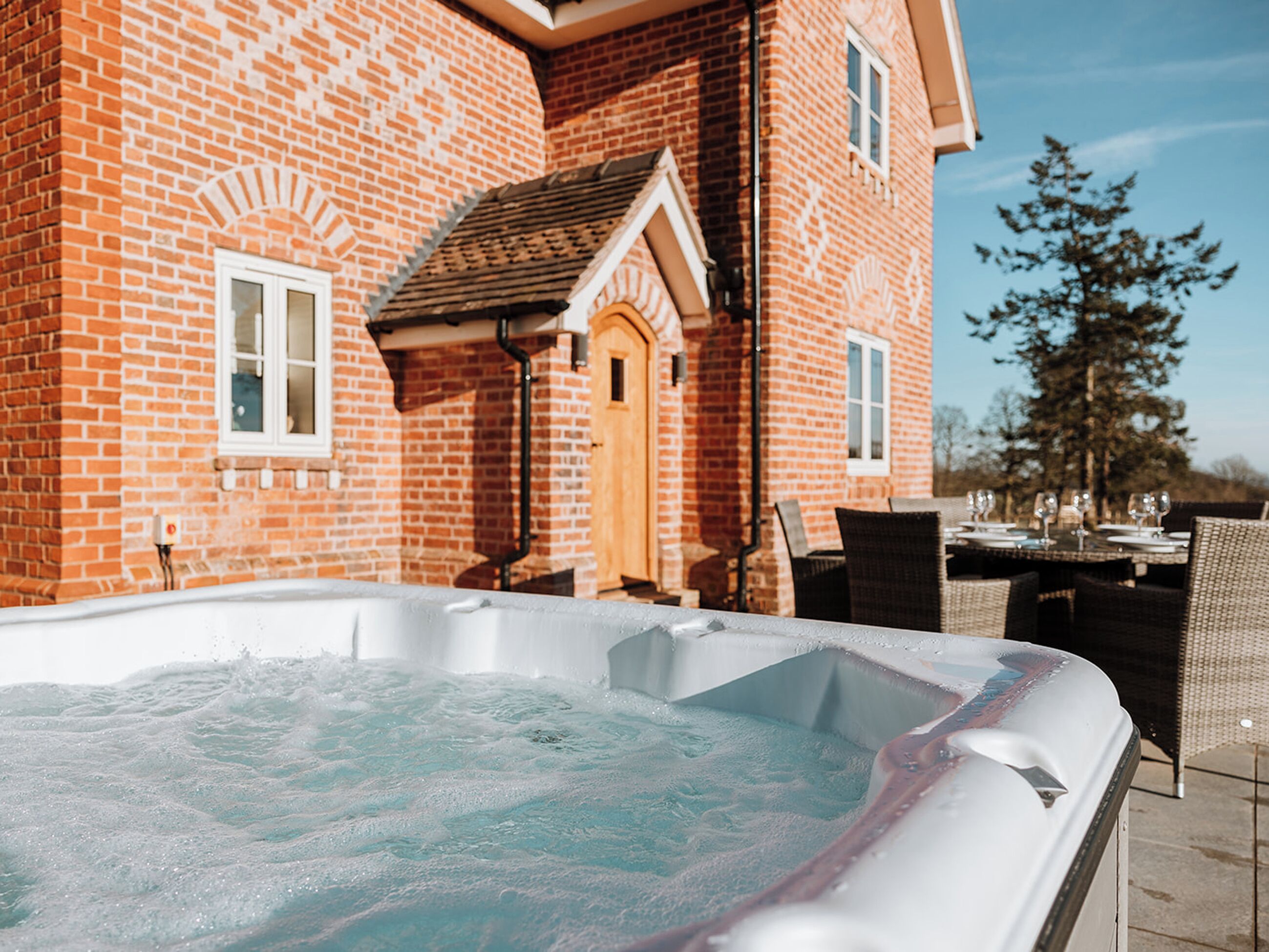A hot tub in front of a brick house with a dining area and outdoor seating.