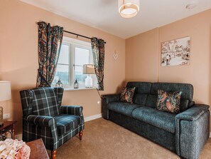 A cozy living room with a plaid armchair, a dark blue sofa, and a window with floral curtains. - Copper Beech Cottage (Welshpool)