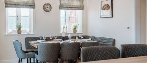A dining area with a round table set for six, surrounded by six grey upholstered chairs. There are two windows with curtains, a wall clock, and a framed picture on the wall.