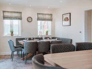 Un coin repas avec une table ronde dressée pour six personnes, entourée de six chaises grises à revêtement rembourré. On y trouve deux fenêtres avec des rideaux, une horloge murale et un tableau encadré accroché au mur.