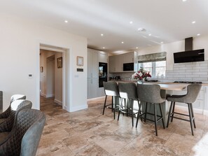A modern kitchen with a central island, bar stools, and a dining area with a cushioned chair. - Copper Beech Cottage (Welshpool)